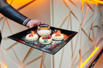 Man holds black square plate with homemade cheese pancakes with fresh berries, honey and sour cream on a beautiful light background with geometric lines. Close-up. Space