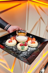 Man holds black square plate with homemade cheese pancakes with fresh berries, honey and sour cream on a beautiful light background with geometric lines. Close-up. Space
