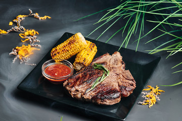 Juicy T-Bone steak with grilled corn and barbecue sauce on a black square plate on a dark smoky...