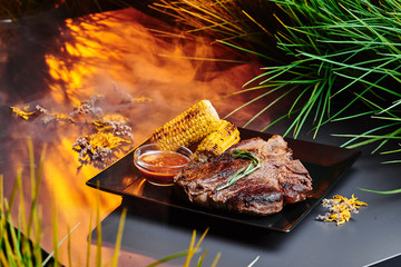 Juicy T-Bone steak with grilled corn and barbecue sauce on a black square plate on a dark smoky...