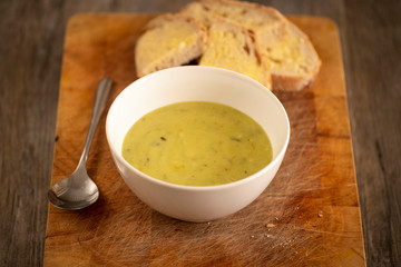 Fresh leek and potato soup in a bowl on a wooden board with crusty white bread and butter