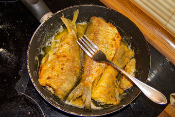 The photo of the whole fried fish without head on the frying-pan or skillet with a fork on the electric cooker