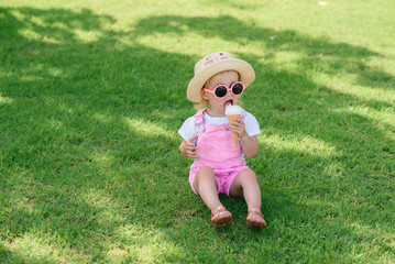 Funny toddler girl wearing pink summer overalls, hat and pink sunglasses sits on a green lawn eats vanilla white ice cream in a sunny garden.