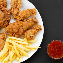 Tasty fried chicken drumsticks, spicy wings, French fries, chicken strips on a white plate over black background, top view. Close-up.