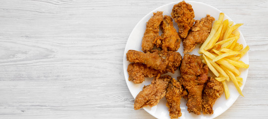 Tasty fried chicken legs, spicy wings, French fries and chicken strips on a white plate over white wooden surface, top view. Flat lay, overhead, from above. Copy space.
