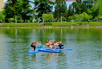 Fototapeta na wymiar Yoga on sup board. Young girls paddling on SUP board on the lake at city. Group women is practicing (doing) yoga, fitness, pilates and meditation on a SUP board. Awesome active training in outdoor. 