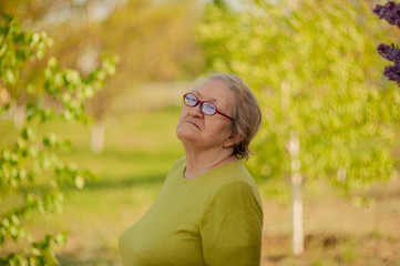 Elderly woman in yellow T-shirt and glasses posing in front of camera against background of bright green birch leaves