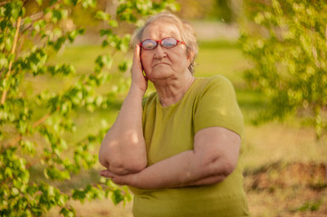 Naklejka na ściany i meble Elderly woman in yellow T-shirt and glasses posing in front of camera against background of bright green birch leaves