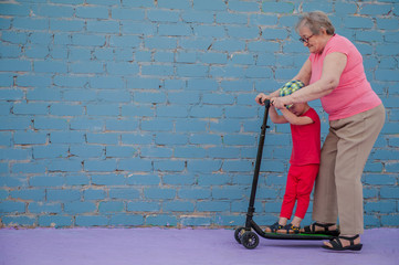 elderly woman in pink T-shirt and glasses with her little grandson is learning to ride new scooter....