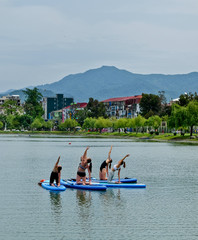 Yoga on sup board. Young girls paddling on SUP board on the lake at city. Group women is practicing (doing) yoga, fitness, pilates and meditation on a SUP board. Awesome active training in outdoor. 