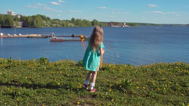 Little blonde girl with blue dress inflates large soap bubbles against backdrop of sea coast. Child plays with transparent balls on sunny day. Concept of summer holidays and outdoor games.