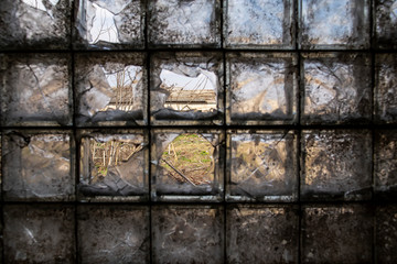 Building of an abandoned and destroyed farm is seen through a partially broken, dirty window of glass blocks in the village. Inside view.