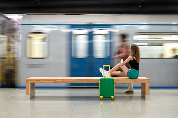 Happy girl traveler waiting for train at metro station sitting on stone bench with legs stretched out on suitcase