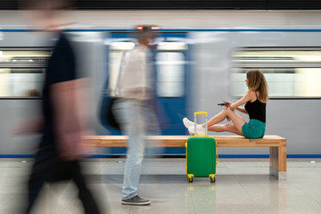 Fototapeta na wymiar a young beautiful european girl with a small green suitcase is sitting on a bench at the railway station waiting for the train. Theme travel alone