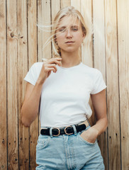 Young pretty girl is standing on a light wooden wall background. Woman is wearing a white empty...