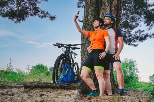 Happy couple with mountain bike taking selfie smartphone outdoors. A happy couple, who lead an active lifestyle, stopped in the woods near the tree to take a photo. Theme sport and active lifestyle