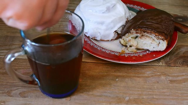 A man is stirring sugar in a cup with tea. Sweet donut and bagel on a plate on wooden table.