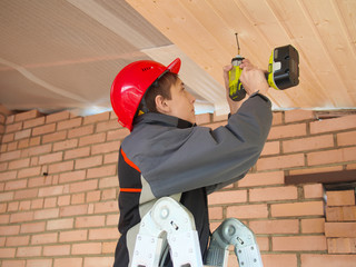 Teenager boy in construction helmet fix clapboard on ceiling of a home with air hammer, nailer and...