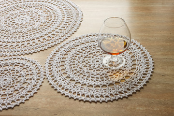 Crocheted round-type napkins of white color, аnd glass of cognac on beige background