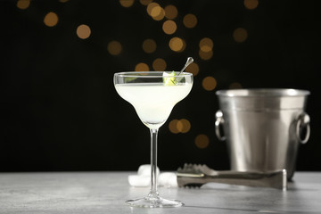 Glass of martini with cucumber, ice bucket and tongs on grey table against blurred lights. Space...