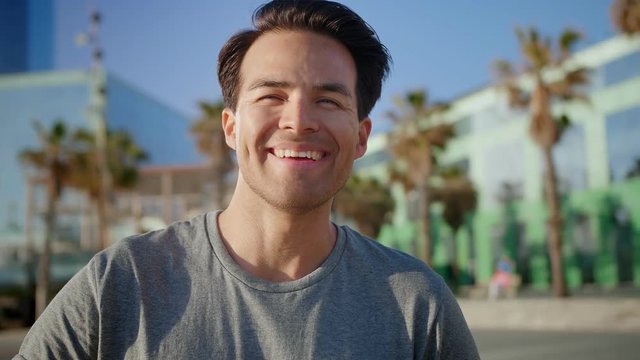 Close portrait of laughing Hispanic healthy adult standing on beach at summer day time, man joyful feeling of vacations and moment of personal happiness standing near glass edification and palms