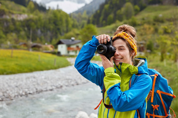 Pleased young female traveler makes photo of mountain and river landscape, poses with rucksack against countryside and forest, pleased to have nice trip, looks at nature horizon catches beautiful view