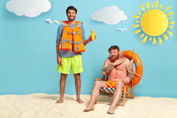 Photo of glad man suggests friend using sunscreen, has positive smile, wears lifejacket, upset red...