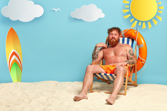 Foxy dissatisfied man gets sunburned at beach, has red skin, sits at sun chair half naked, has tattooed arms, life ring and surfboard, white clouds needs lotion for body. Sun skin care concept