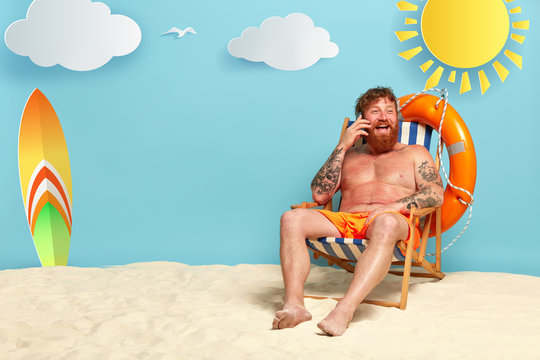 Image of happy redhead man in shorts glad to have telephone conversation with friend, has red sunburnt skin, tells about vacation at resort place, laughs positively, expresses good emotions.