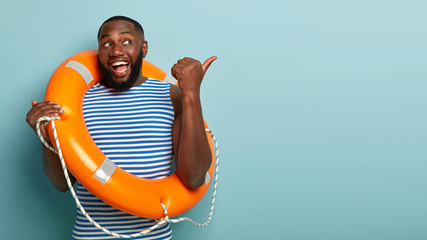 Positive dark skinned man with thick bristle, muscular body, has glad expression, points away on...