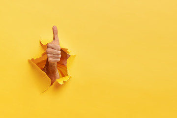 Unrecognizable man shows like gesture through torn yellow wall, keeps thumb up, says you are best,...