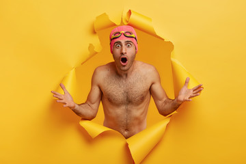 Shocked embarrassed naked man spreads hand, has naked body, keeps mouth wide opened, wears swimcap and goggles, cannot believe in his failure on swimming competiton, stands in ripped background - obrazy, fototapety, plakaty
