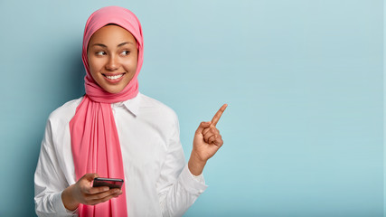 Joyful pleasant looking young Muslim woman with dark skin points at upper right corner, holds smartphone device, shows free space for your advertising content, wears pink hijab, white shirt. Look here