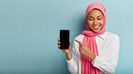 Positive lovely young woman with pleasant smile, points at blank screen of cellular, has happy...