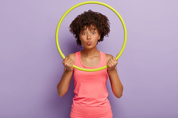 Athlete funny dark skinned woman looks through hula hoop, has folded lips, burns excess fat in body, strengthens muscles, increases blood flow wears casual pink vest models indoor with sport equipment