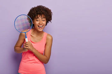 Happy tennis girl holds racket, glad to win game, wears casual pink vest, stands against purple background with free space. Fitness woman enjoys awesome tennis tournament. Recreation concept