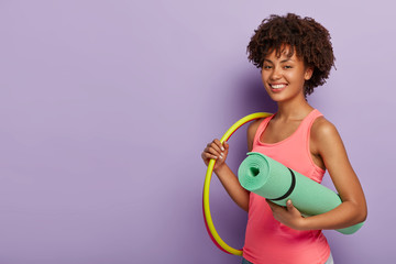 Delighted healthy woman rotates hoop, carries rolled karemat, has training, leads healthy lifestyle, ready to have fitness lessons or gymnastics, isolated over purple wall, attends sport section