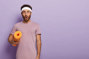 Emotional guy with bristle wears headband and purple t shirt, gets ready for aerobics, holds karemat, stares at camera, isolated over purple background with free space for your text information