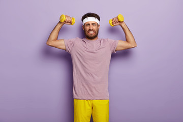 Determined young sporty unshaven man clenches teeth, raises muscular arms, does exercises with dumbbells, clenches teeth, wears white headband, t shirt and shorts, isolated over purple background.