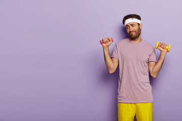 Young man looks unwillingly, holds dumbbells in both hands, has no desire for training, dresses in active wear, stands against purple background with copy space for your advertising content.
