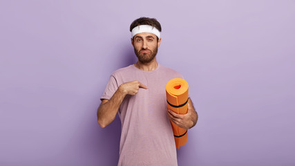 You mean me? Confused unshaven guy points at himself, wears white headband, dressed casually, holds yellow fitness mat, asks whether he should conduct workout training, isolated on purple wall