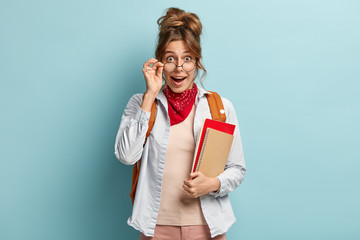 Surprised happy female student looks through glasses, holds hand on frame, carries spiral notebook...