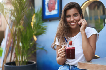 Alluring cute flirty young girl tanned brunette, touch cheek joyfully smiling hold strawberry shake smoothie listen interesting story look coquettish have romantic date outdoor cafe street terrace