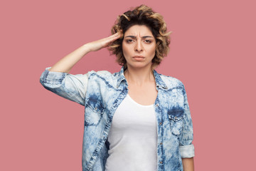 Obraz premium Yes sir. Portrait of serious young worker woman with curly hairstyle in casual blue shirt standing and looking at camera with salute and ready to do smoething. studio shot, isolated on pink background