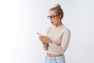 Woman browsing internet via smartphone gasping shocked and stunned holding mobile phone reacting...