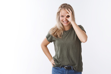 Charismatic young happy woman with blond hair and blue eyes peeking through hole showing okay gesture on eye and gazing at camera amused and joyful grinning cheeky against white background