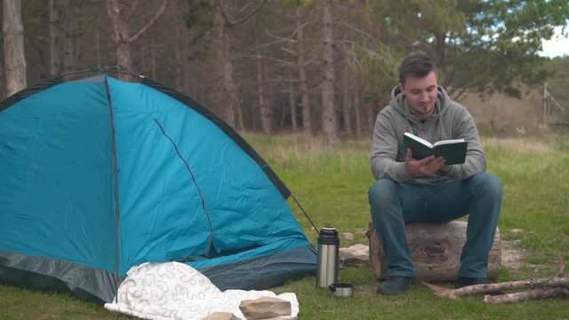 A young guy sits on a large log near a blue tent and Temos in the forest and reads a book. Background - pine forest.