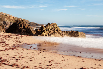 Praia com as algas vermelhas na areia trazidas pelas ondas que rebentam na areia com as rochas ao fundo do areal. Praia situada a sul de Sines em Portugal, Europa.