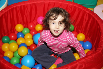 Fototapeta na wymiar Little curly cute smile girl plays in balls for a dry pool. Play room. Happiness.