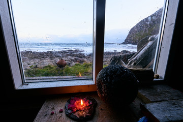 Look through the window of a cabin at the coast with a burning candle in the foreground.